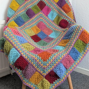 May include: A colorful, handmade crochet blanket draped over a chair. The blanket features a patchwork design with squares in shades of yellow, green, blue, pink, orange, and purple. The edges are finished with a contrasting border.