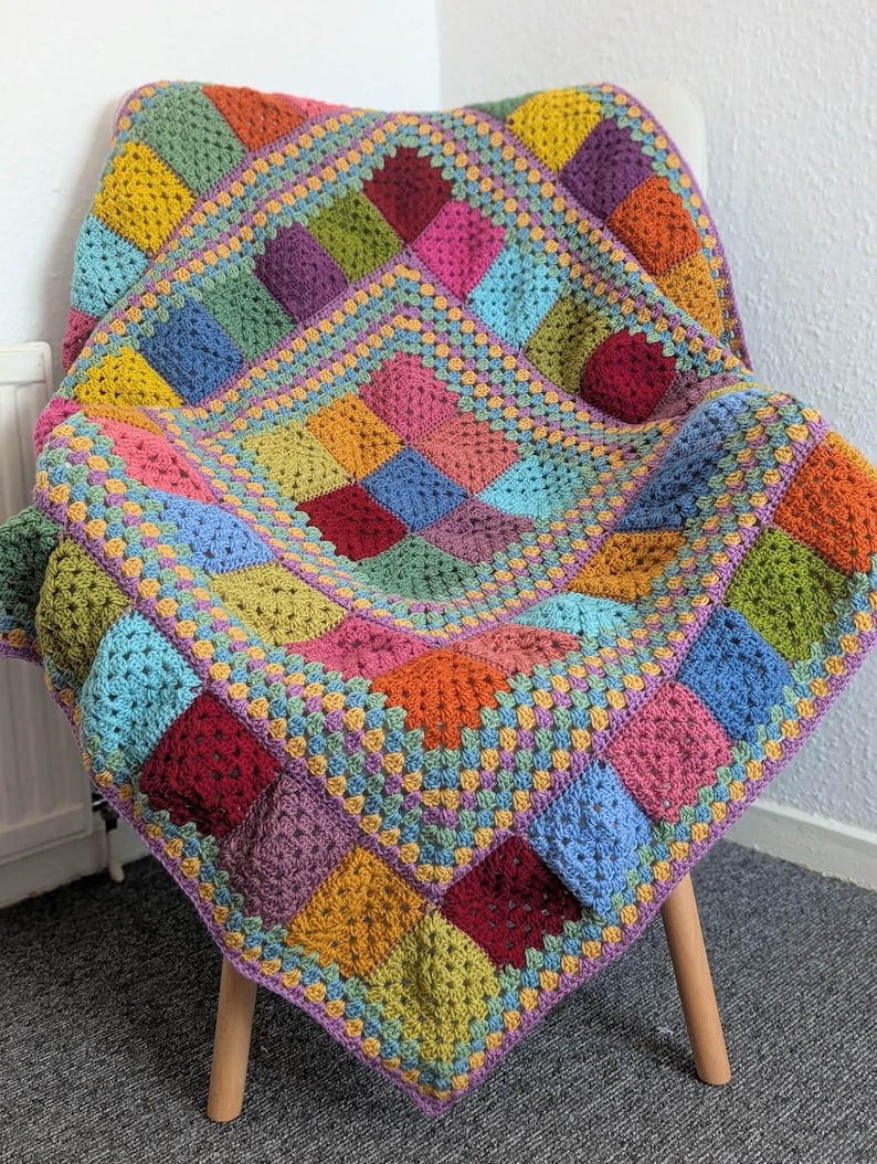 May include: A colorful, handmade crochet blanket draped over a chair. The blanket features a patchwork design with squares in shades of yellow, green, blue, pink, orange, and purple. The edges are finished with a contrasting border.