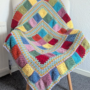 May include: A colorful, handmade crochet blanket draped over a white chair. The blanket features a patchwork design with squares in various colors, including red, yellow, blue, green, and pink. The blanket is approximately 50 inches by 50 inches.