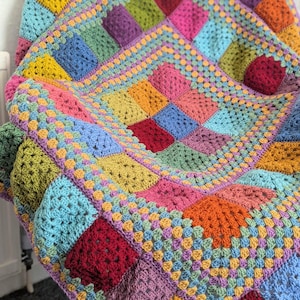 May include: A colorful, handmade crochet blanket. The blanket features a patchwork design with squares in various colors, including yellow, blue, green, pink, and purple. The blanket is draped over a chair, showcasing its texture and design.