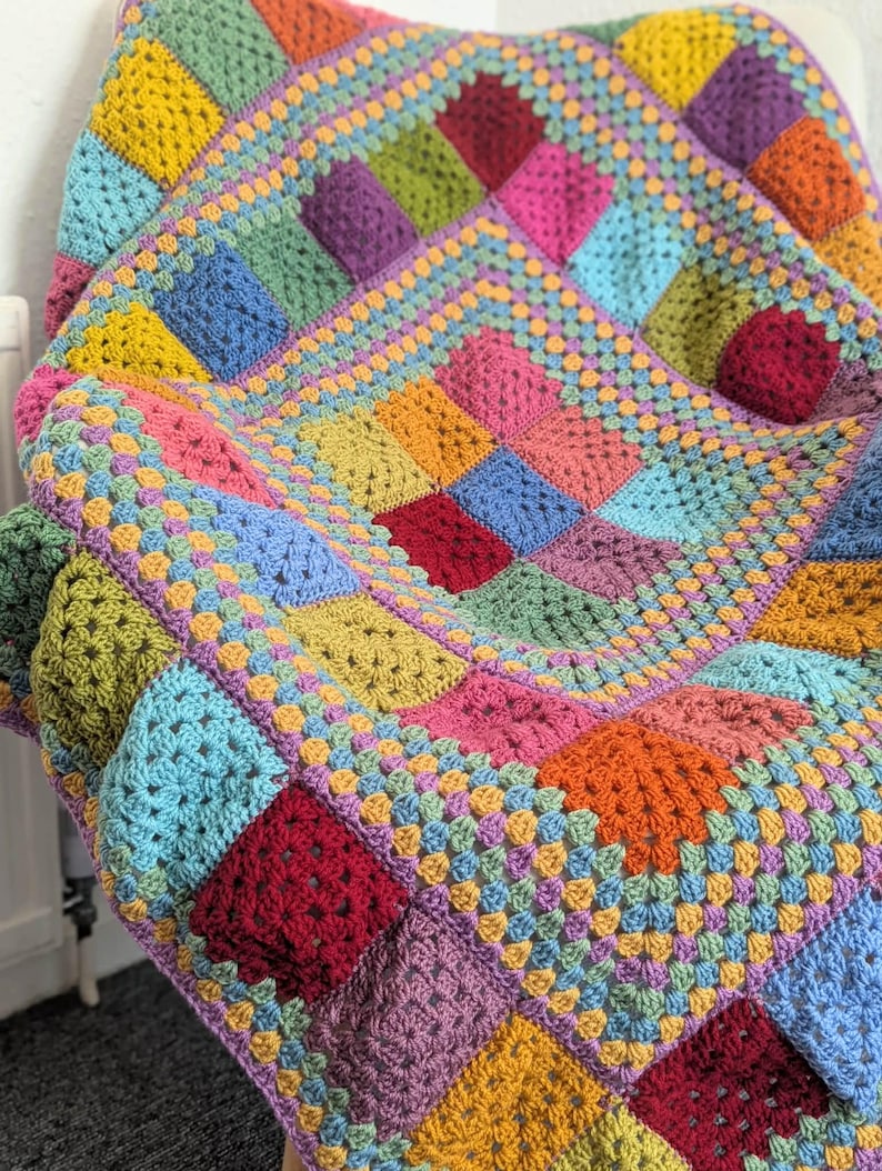 May include: A colorful, handmade crochet blanket. The blanket features a patchwork design with squares in various colors, including yellow, blue, green, pink, and purple. The blanket is draped over a chair, showcasing its texture and design.