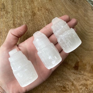 May include: Three white selenite crystal towers, each with a unique shape and texture, are displayed on a wooden surface.