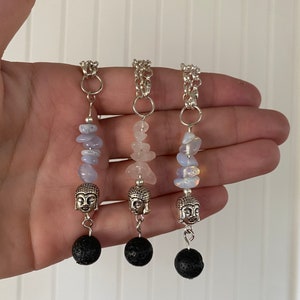 Puede incluir: Tres colgantes de cristal hechos a mano con cadenas de plata. Cada colgante presenta una cabeza de Buda plateada, una cuenta de lava negra y un grupo de piedras preciosas azules, rosas y blancas. Los colgantes se muestran sobre un fondo blanco.