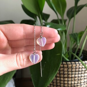 May include: A pair of silver chain earrings with iridescent heart-shaped charms. The charms are a light purple color.
