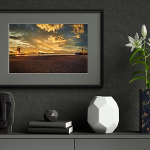 May include: A framed photograph of a sunset over a beach with a city skyline in the distance. The photo is displayed on a shelf with a black sculpture, a geometric white vase, and a vase with white flowers.
