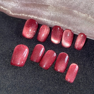 May include: A set of ten oval-shaped, glossy, red press-on nails. The nails have a shimmering, glittery effect, with a darker shade at the base that fades to a lighter, more sparkly tip. The nails are arranged on a black surface.