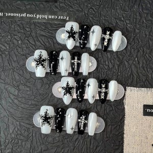 May include: A set of twenty press-on nails in a coffin shape, featuring a black and white color palette. The designs include black star charms, silver cross embellishments, and hand-painted silver star accents on glossy black and white surfaces.