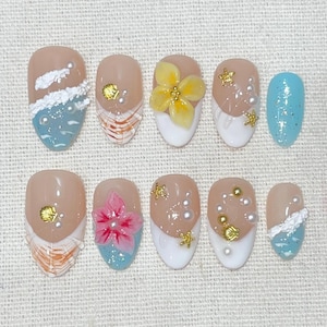 May include: A set of ten artificial nails with a variety of designs. The nails feature a nude base with white, blue, and gold accents. Some nails have 3D floral and seashell embellishments, while others have pearl accents.