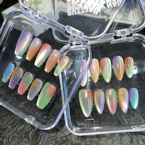 Op de afbeelding: Twee doorzichtige plastic doosjes gevuld met iriserende, ovaalvormige valse nagels. De nagels vertonen een kleurengradatie, waaronder paars, oranje, groen en blauw, waardoor een glinsterend effect ontstaat. De doosjes zijn open en onthullen de nagelsets.