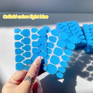 May include: Multiple sheets of light blue adhesive dots, designed for holding items in place. The adhesive dots are arranged in a grid pattern on each sheet. The sheets are held in a hand.