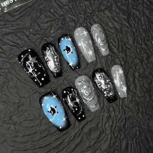 May include: A set of ten press-on nails featuring a celestial theme with black, light blue, and grey base colors. The designs include white star motifs, silver metallic swirls, and raised silver spheres on a glossy finish with coffin-shaped tips.