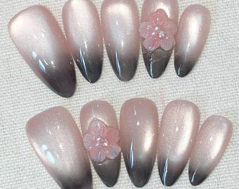 May include: A set of ten oval-shaped artificial nails. Each nail features a gradient design, transitioning from a light pink hue to a dark gray at the tips. Two nails are adorned with pink flower embellishments.