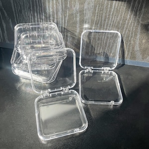 May include: A stack of clear plastic square boxes with hinged lids. The boxes are empty and are arranged on a dark gray surface.
