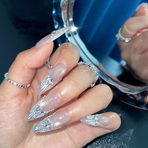 Può includere: Primo piano di una mano con unghie lunghe, trasparenti e appuntite decorate con glitter argentato e strass.