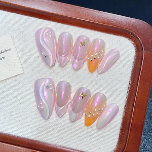 Könnte beinhalten: Ein Set aus schillernden rosa und orangefarbenen Kunstnägeln. Einige Nägel weisen Perlen- und Strassakzente auf, während andere goldene Sternverzierungen haben. Die Nägel sind auf einer weißen Oberfläche angeordnet.