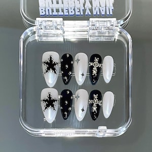 May include: A set of press-on nails in a clear plastic case. The nails feature a variety of designs, including a star, small studs, a starburst, and a cross, all in silver. The nails are white and black.
