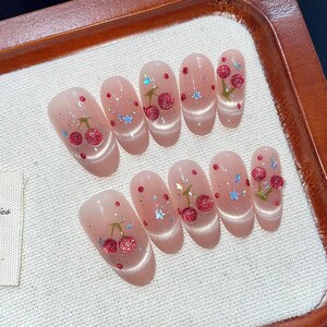 Puede incluir: Conjunto de diez u&ntilde;as postizas ovaladas con base rosa y dise&ntilde;o de cereza. Cada u&ntilde;a tiene una cereza con tallo verde y fruta roja, y algunas tienen detalles de purpurina.