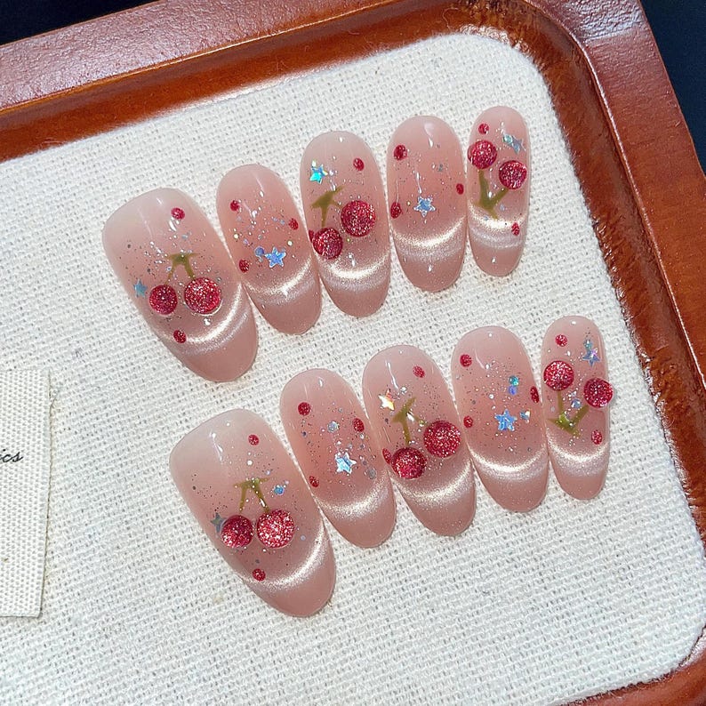 Puede incluir: Conjunto de diez u&ntilde;as postizas ovaladas con base rosa y dise&ntilde;o de cereza. Cada u&ntilde;a tiene una cereza con tallo verde y fruta roja, y algunas tienen detalles de purpurina.