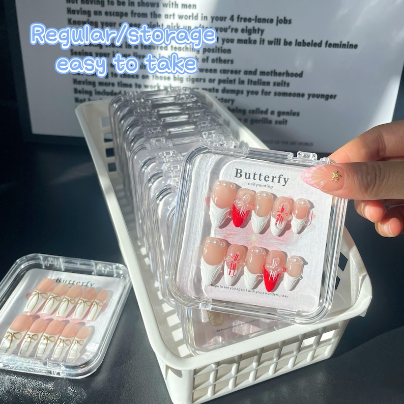 May include: A clear plastic storage box with multiple compartments, each containing a set of fake nails. The nails are white with red and white designs. The box has the word "Butterfly" printed on it.