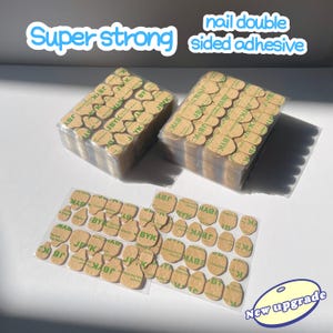 Könnte beinhalten: Nahaufnahme einer Packung superstarker doppelseitiger Klebepads für Nägel. Die Packung ist mit "Super strong" und "nail double sided adhesive" beschriftet. Die Pads sind klein, rund und beige mit einem grünen Etikett. Die Packung befindet sich auf einer weißen Oberfläche. Im Vordergrund befindet sich eine kleinere Packung der gleichen Pads. Der Text "New upgrade" ist in einem blauen Kreis auf der rechten Seite des Bildes geschrieben.