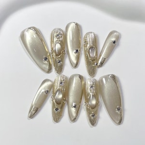 Puede incluir: Conjunto de diez uñas postizas blancas y doradas con acabado perlado y detalles decorativos. Cada uña presenta un diseño dorado con un pequeño strass transparente.