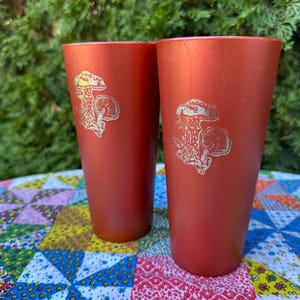 May include: Two tall, orange-red tumblers with a silver mushroom design. The tumblers are sitting on a colorful patchwork quilt.