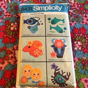 May include: A vintage Simplicity transfer pattern packet featuring zodiac signs: Aquarius, Pisces, Aries, Taurus, Gemini, and Cancer. The packet has a blue and white design with the number 8431. The background is a colorful floral pattern.
