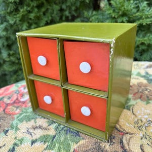 May include: A square olive green storage unit featuring four individual drawers with bright orange fronts. Each drawer is equipped with a small, round, white knob centered on the face. The unit has a smooth finish with visible wear along the right edge.