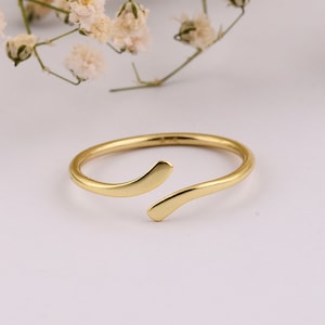 May include: A gold-colored adjustable ring with an open design. The ring features a smooth, curved band that wraps around the finger, with two ends that gently overlap. The ring is set against a soft, white background with delicate floral accents.