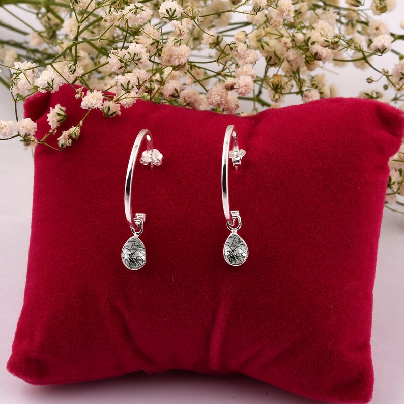 May include: A pair of silver hoop earrings with a teardrop-shaped crystal pendant. The earrings are displayed on a red velvet cushion.