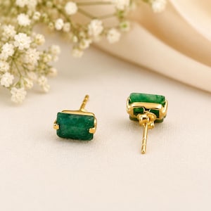 Puede incluir: Un par de pendientes de botón con piedras rectangulares de color verde intenso engastadas en metal dorado pulido. Un pendiente está colocado para mostrar la cara frontal de la piedra verde, mientras que el otro está girado para mostrar el poste.