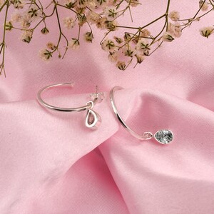 May include: A pair of silver hoop earrings with a teardrop-shaped crystal pendant hanging from each hoop. The earrings are on a pink fabric background.