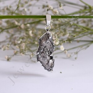May include: A silver pendant featuring a raw, dark grey meteorite. The pendant is suspended from a silver bail.