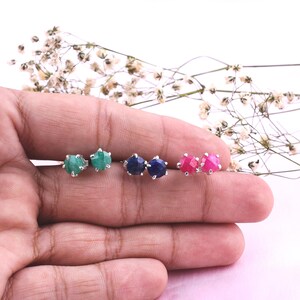 Puede incluir: Tres pares de pendientes de botón con piedras preciosas redondas y facetadas en verde, azul oscuro y rosa brillante. Cada piedra está engastada en una montura de metal plateado con garras, creando una colección de joyas circulares pequeñas.