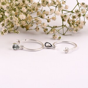 May include: A pair of silver hoop earrings with a teardrop-shaped gemstone pendant. The gemstone is a light green color with a sparkly finish.
