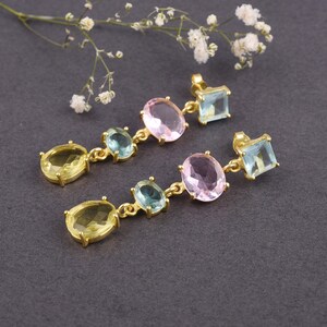 Pode incluir: Um par de brincos dourados com um design linear. Cada brinco apresenta uma pedra oval amarela, uma pedra oval azul-esverdeada e uma pedra oval rosa, conectadas por uma delicada corrente. Uma pedra azul-esverdeada quadrada completa o design.