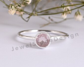 May include: A delicate silver ring featuring a round, light pink gemstone. The ring has a thin band and a bezel setting that holds the gemstone securely. The design is simple and elegant, suitable for everyday wear.