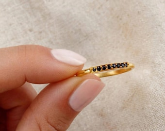 May include: A thin gold band ring featuring a horizontal row of seven small, round, black gemstones set into a flat, rectangular metal bar section. The ring has a polished metallic finish and a simple, slender design.