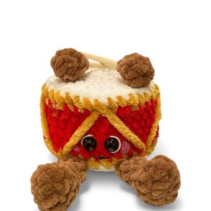 Puede incluir: Un juguete de tambor de peluche hecho a mano con una cara sonriente, ojos negros grandes y baquetas marrones y esponjosas. El tambor es rojo con adornos dorados y pies marrones y esponjosos. La parte superior es blanca.
