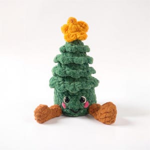 Könnte beinhalten: Ein handgehäkelter, grüner Weihnachtsbaum mit einem gelben Stern. Der Baum hat ein lächelndes Gesicht, rosige Wangen und braune Arme und Füße. Eine skurrile Weihnachtsdekoration für die Feiertage.