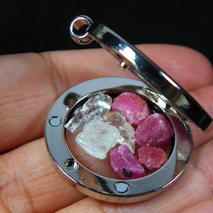 Peut inclure: Un pendentif en argent avec un médaillon en verre transparent, révélant une collection de petites pierres précieuses roses et blanches. Le médaillon a un couvercle à charnière et une fermeture magnétique.