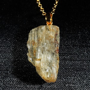Puede incluir: Un colgante de cristal en bruto, translúcido y de color amarillo pálido, suspendido de una cadena de metal dorado. La piedra presenta una textura estriada vertical con sutiles inclusiones de color marrón rojizo en su forma irregular, alargada y dentada.