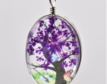 Puede incluir: Un colgante de plata con un dije de resina ovalado transparente que contiene un arreglo de flores moradas prensadas. El arreglo floral se asemeja a un árbol con un tronco y ramas de color marrón oscuro.