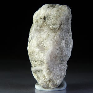 May include: A light-coloured, rough rock with a textured surface. The rock displays a mix of grey, white, and pale purple hues. It is set against a dark background, highlighting its natural form and details.