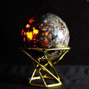 Op de afbeelding: Een gepolijste, bolvormige steen met een gevlekte grijze, zwarte en bruine oppervlaktestructuur, met een helder, gloeiend oranje licht dat aan één kant van binnenuit straalt. De bol rust op een geometrische, goudkleurige metalen draadstandaard.
