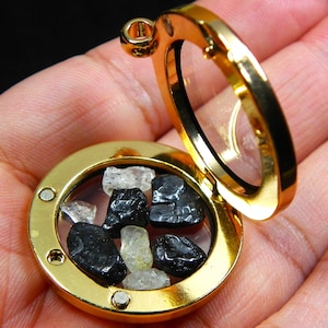 May include: A gold-coloured, round locket with a hinged opening, filled with black and clear gemstones. The locket is open, revealing the stones inside. The background is a hand.