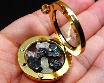 May include: A gold-colored, round locket with a hinged opening, filled with black and clear gemstones. The locket is open, revealing the stones inside. The background is a hand.
