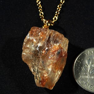 May include: A raw, translucent amber-toned gemstone pendant suspended from a gold-coloured chain, positioned next to a United States dime for scale, showcasing natural irregular facets and internal reddish-brown mineral inclusions.
