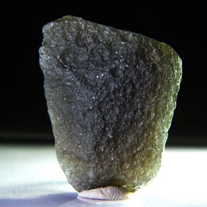 May include: A rough, irregularly shaped, olive-green gemstone with a textured surface. The stone has a dark, almost black, appearance in some areas, with lighter green highlights. The gemstone is set against a dark background.