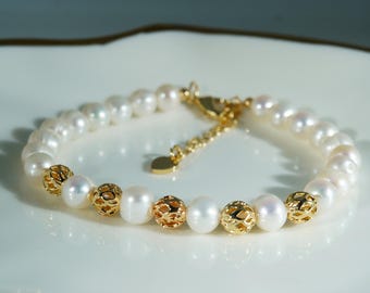 Könnte beinhalten: Ein Armband mit einer Reihe weißer, runder Perlen, die mit goldenen, kugelförmigen Filigranperlen durchsetzt sind. Das Schmuckstück enthält eine goldfarbene Metallkettenverlängerung mit einem kleinen, flachen, herzförmigen Anhänger am Karabiner.